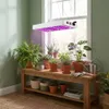 A grow light illuminates a collection of potted plants on a wooden bench in front of a window. Náhľad obrázku produktu