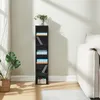 A tall, narrow black shelf unit stands against a light gray wall, with a vase of flowers on top and books and CDs on its shelves. A large green plant in a woven basket sits to the left of the shelf, and part of a cream-colored sofa is visible to the right. Náhled obrázku produktu