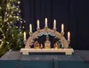 A wooden Christmas arch with illuminated houses and candle-shaped lights, set against a dark background and a decorated Christmas tree. Podgląd zdjęcia produktu