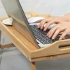 A person's hands are typing on a laptop placed on a bamboo lap desk. A white computer mouse and a small white dish are also on the desk. Produktbild-Vorschau