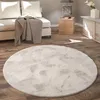 A round, fluffy rug is placed on a wooden floor in a living room setting, with a sofa and side table visible. Produktbild-Vorschau