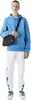 A man wearing a blue hoodie and white pants with blue accents, holding a black bag. Voir les images du produit
