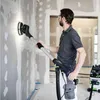 A man is sanding a wall with a long-handled sander. There is a vacuum cleaner in the background. Náhled obrázku produktu