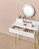 A white vanity desk with gold legs, featuring a lighted mirror and organized makeup in its drawers. The desk is placed against a pink wall with a wooden floor. Produktbild-Vorschau
