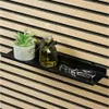 A black shelf mounted on a slatted wooden wall, displaying a small potted plant, a bottle of liquid with a black ribbon, and a set of keys. Produktbild-Vorschau