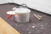 Painting supplies are laid out on a protective floor covering, with paint splatters visible on the floor. Produktbild-Vorschau