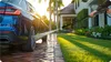 A person is using a pressure washer to clean a driveway next to a car and a lawn. Produktbild-Vorschau