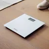 A white digital bathroom scale is placed on a wooden floor next to a rug and a slipper. Podgląd zdjęcia produktu