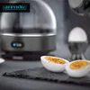 A close-up of a modern egg cooker with two halved hard-boiled eggs in the foreground, seasoned with pepper. Produktbild-Vorschau