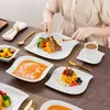 A person is eating a meal served on white, leaf-shaped plates, with various dishes including steak, soup, and fruit salad. Produktbild-Vorschau