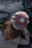 A person wearing a colorful crocheted hat, seen from behind, with a stone wall in the background. Produktbild-Vorschau