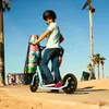 A young person rides a blue kick scooter on a sunny day near a beach with palm trees and graffiti art. Anteprima dell’immagine prodotto