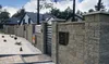 A black mailbox is mounted on a stone wall in front of a modern house and fence. Produktbild-Vorschau