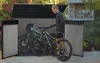 A man is putting two bicycles into a large outdoor storage shed. Anteprima dell’immagine prodotto