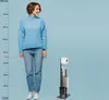 A woman stands next to a toilet paper holder and brush stand, with a height chart on the left. Voir les images du produit