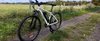 An electric bicycle is parked on a dirt path next to a grassy field with a fence. Produktbild-Vorschau