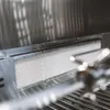 Close-up view of the interior of a stainless steel grill, showing a ceramic burner cover and grill grates. Náhľad obrázku produktu