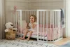 A young girl is sitting in a white crib with pink bedding. Produktbild-Vorschau