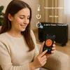 A woman is holding a smartphone and interacting with an app, with a black electric heater visible in the background. Icons and text indicating temperature selection, on/off, and timer functions are overlaid on the image. Náhľad obrázku produktu