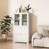 A white wooden cabinet with glass doors and drawers, placed in a room next to a sofa and a plant. Podgląd zdjęcia produktu