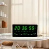 A digital clock with a calendar and temperature display sits on a glass table in a well-lit room. Produktbild-Vorschau