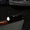 A black flashlight with its light on, resting on the edge of a white boat. The background is dark and out of focus, showing other boats and a pebbly shore. Náhled obrázku produktu