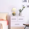 A bedside table with a lamp and a vase of greenery next to a bed. Náhľad obrázku produktu