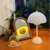 A white mushroom-shaped lamp sits on a wooden surface next to a small plant and a mirror reflecting the lamp's light. Produktbild-Vorschau