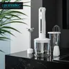 A hand blender set is displayed on a dark marble countertop, with a large green plant in the background. Produktbild-Vorschau