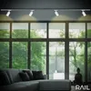 A living room scene with a sectional sofa, large windows with a view of trees, and track lighting. Produktbild-Vorschau