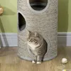 A cat sits inside a multi-level cat scratching tower with a ball of yarn on the floor. Produktbild-Vorschau
