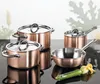 A set of copper and stainless steel cookware is displayed on a reflective black surface against a blurred background. Produktbild-Vorschau
