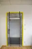 A doorway covered with a clear plastic sheet, framed by yellow tape, with black straps holding the plastic in place. A fire extinguisher is visible on the left side. Produktbild-Vorschau