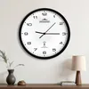 A wall clock is mounted on a white wall above a wooden shelf with a vase and a lamp. Produktbild-Vorschau