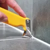 A hand is using a yellow tool to remove sealant from a corner where two tiled surfaces meet. Produktbild-Vorschau