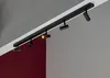 A black track lighting system mounted on a ceiling, with a dark red wall below. Náhľad obrázku produktu