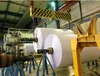 Large rolls of white paper are displayed in an industrial setting, possibly a paper mill or printing facility. A crane is visible overhead, and machinery is present in the background. Anteprima dell’immagine prodotto