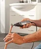 A person is using a Dremel rotary tool to cut a copper pipe in a bathroom setting. Produktbild-Vorschau