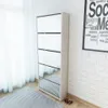 A tall, white shoe cabinet with mirrored doors stands against a white brick wall next to a sheer white curtain. A striped rug is on the floor in front of the cabinet, and a pair of red high-heeled shoes are on the floor to the right of the cabinet. Náhľad obrázku produktu