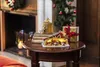 A Christmas nativity scene is displayed on a wooden table, with a fireplace and Christmas tree in the background. Podgląd zdjęcia produktu