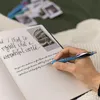 A hand is writing in a journal with a blue pen. There are photos and other pens in the background. Podgląd zdjęcia produktu