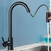 A black kitchen faucet with a pull-down sprayer is shown in use, with water flowing into a sink. Two glasses with blue liquid are visible in the background. Produktbild-Vorschau