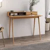 A wooden desk with a shelf and drawer, set against a white wall with a grey rug and a basket in the background. Produktbild-Vorschau