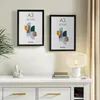 Two black A3 picture frames are displayed on a wall above a white dresser with a lamp and books on top. Produktbild-Vorschau