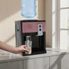 A person is filling a glass with water from a water dispenser in a home or office setting. Náhled obrázku produktu
