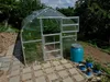 A greenhouse in a garden setting with a blue water barrel and potted plants. Produktbild-Vorschau