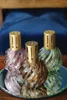 Three decorative glass bottles with gold caps are arranged on a reflective surface, with a blurred blue upholstered headboard in the background. Produktbild-Vorschau