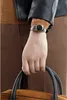 A close-up shot of a person's hand wearing a watch and holding a bag. Produktbild-Vorschau