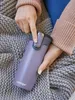 A person is holding a purple Contigo travel mug while sitting on a gray knitted blanket. Produktbild-Vorschau