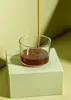 A glass of amber liquid is being poured into from a thin stream, sitting on a light green box against a light green and yellow background. Produktbild-Vorschau